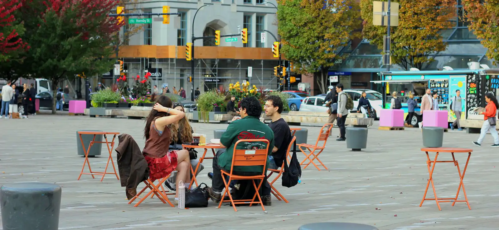 People sitting at orange café tables in a public plaza in downtown Vancouver on an autumn day